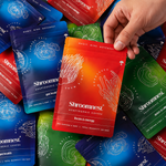 A hand holds a Shroomnest Marigold Coffee pouch in the 'Focus & Energy' flavor, which features a red-to-orange gradient. Multiple other colorful Shroomnest coffee pouches in blue, green, and red are scattered in the background.